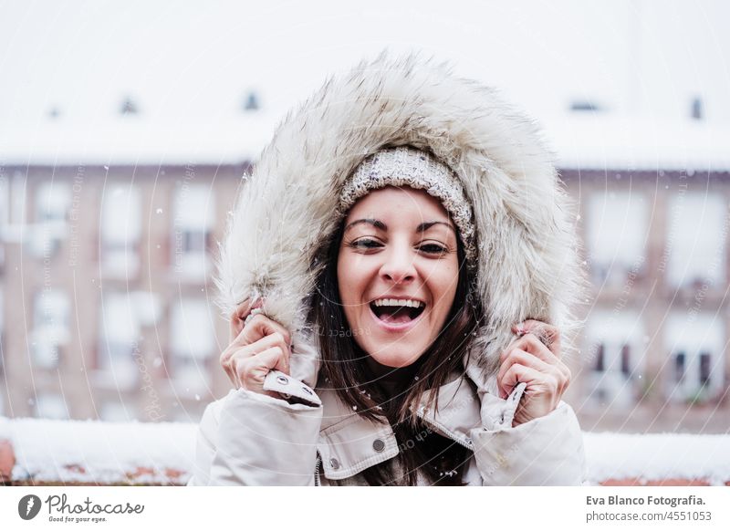 happy woman on terrace at home enjoying snow holding hood with hands, neighborhood background. Snow in the city caucasian urban snowing snow flake smiling 30s