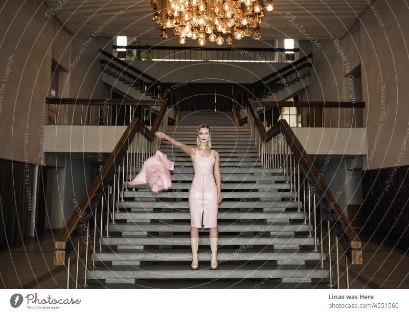A vintage mansion is a perfect place for a gorgeous fashion model to show a pink latex outfit and a pretty face. Massive chandelier, retro stairs, pink outfit. The main subjects of this scene.