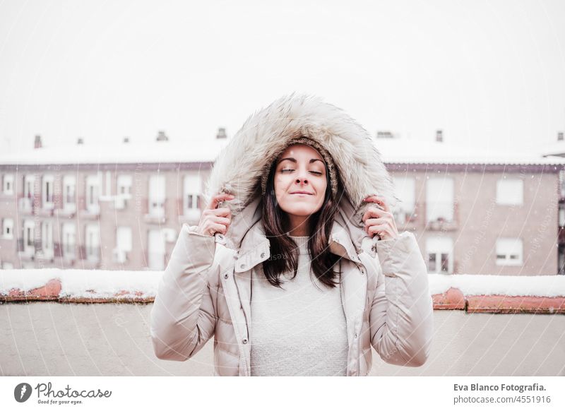 relaxed woman with eyes closed on terrace at home enjoying snow holding hood with hands, neighborhood background. Snow in the city winter portrait caucasian