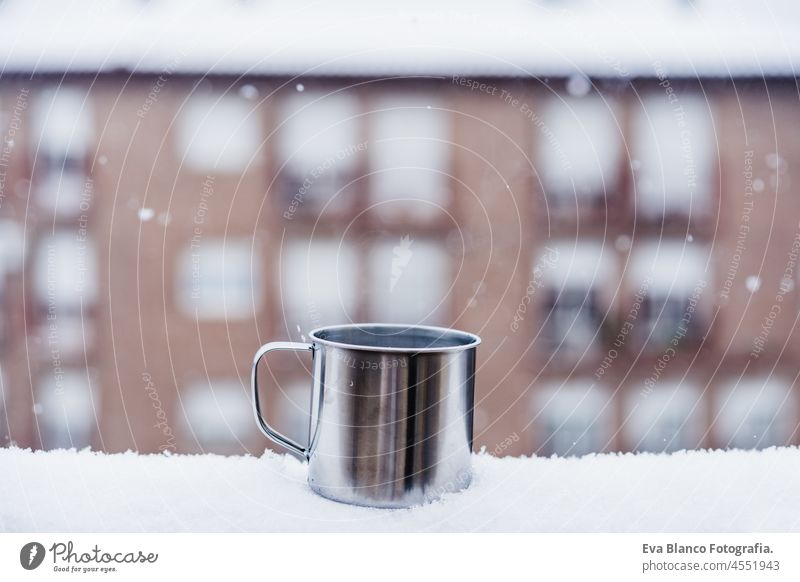metallic cup of hot drink, tea or coffee, on terrace during snow in winter, neighborhood background. Snow in the city. Nobody nobody home urban white cold