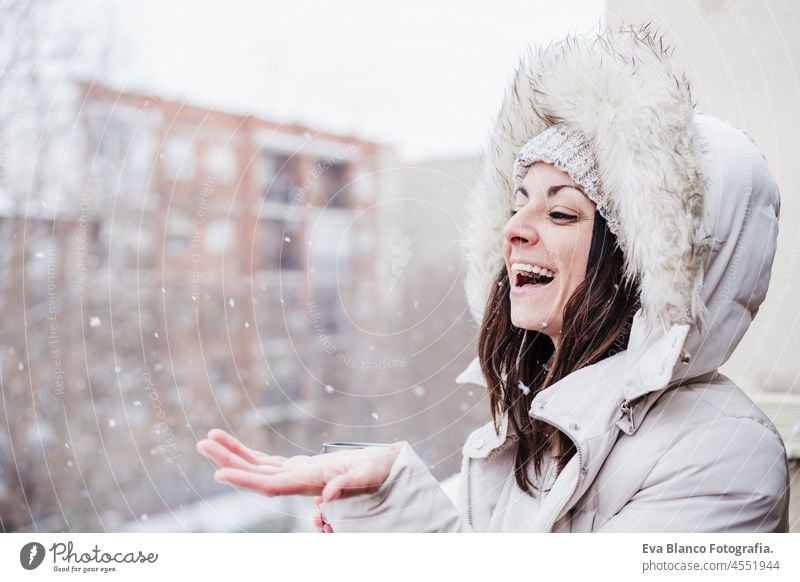 happy woman on terrace at home enjoying snow holding hood with hands, neighborhood background. Snow in the city winter caucasian smiling white snowing