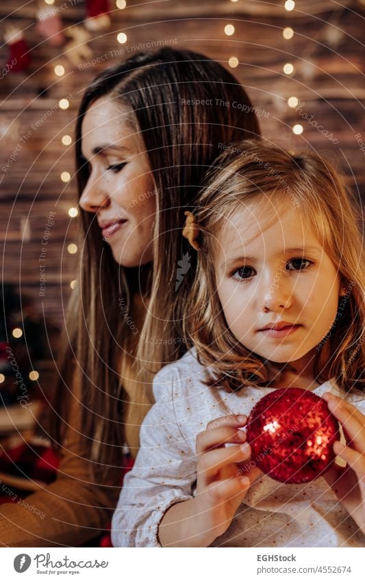 Hispanic mom holding her daughter with red ball and lights. . Living room decorated by Christmas tree and present gift box, the light give cozy atmosphere. New Year and mummy and daughter concept