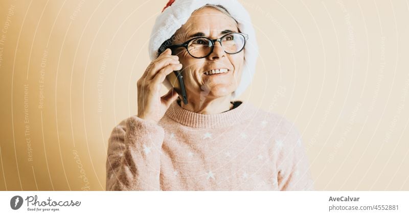 Happy Joyful senior old white hair woman wearing santa claus hat smiling while using smart phone. stands against a color background. Wearing Christmas clothes. Copy space. Advertising space