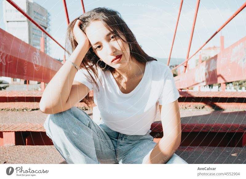Urban shot of a modeling young african woman with long hair sit on a red floor looking serious to camera. City urban concept. Happy day on the city, white shirt blue jeans. Modern outfit styling.