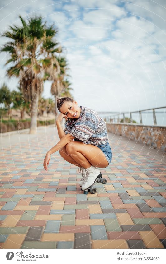 Positive woman in roller skates squatting on walkway street sportive hobby sidewalk activity training wellbeing healthy lifestyle female footwear fit workout