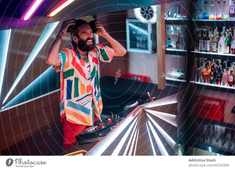 Joyful young disc jockey laughing during party in club man dj mix music console nightlife nightclub cheerful controller modern male beard shirt headphones