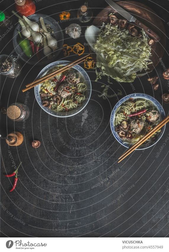 Food background with traditional asian soup in bowls with chopsticks, cabbage, garlic, chili and shiitake on dark concrete table. Spicy ramen with healthy vegetables at home. Top view with copy space.