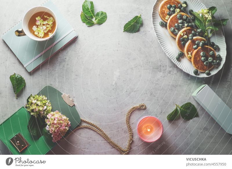 Female freelancer background with breakfast and cosmetic. Pancakes with blueberries, herbal tea, candle, hydrangea, green handbag and notebook on pale concrete table. Top view with copy space.