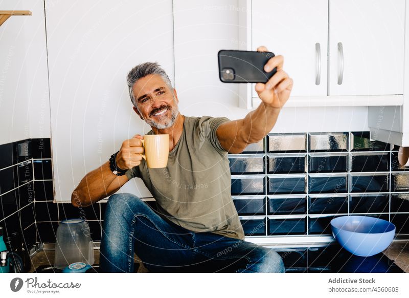 Bearded senior male taking selfie in kitchen man cellphone at home smartphone leisure lifestyle counter drink cup tea handsome chill memory mobile rest