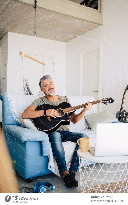 Elderly guitarist performing music on sofa near laptop man musician loft instrument sign interior acoustic play online headphones chill couch meloman aged hobby