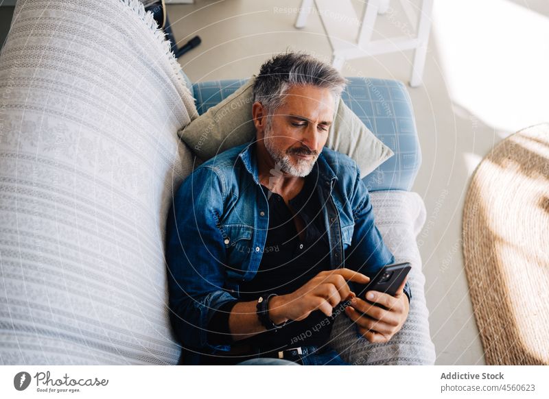 Senior man browsing cellphone lying down on couch smartphone mobile sofa using style loft chill rest pastime living room aged jeans modern free time denim relax