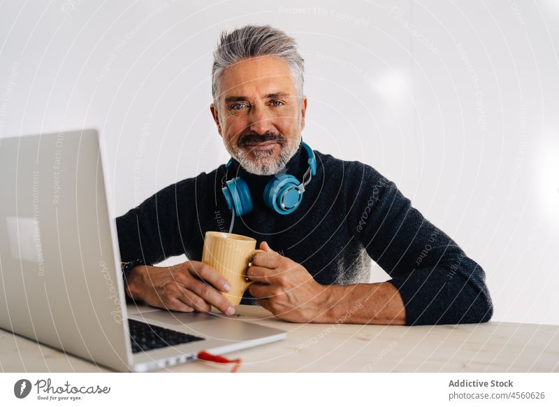 Aged blogger resting with cup of tea in studio man drink podcast broadcast microphone laptop record workspace mug break headphones vlog elderly senior table