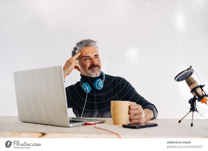 Aged blogger resting with cup of tea in studio man drink podcast broadcast microphone laptop record workspace mug break headphones vlog elderly senior table