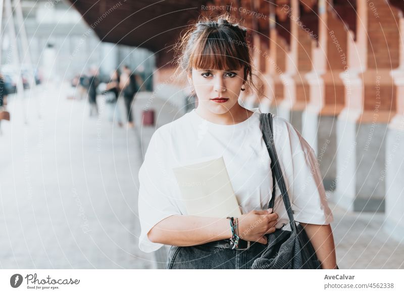 Young beautiful red head looking serious to camera hipster woman .Holding a book near the train station. Trendy girl in summer T-shirt .pensive female posing in the street near a brick wall.