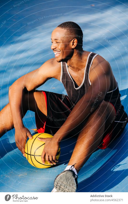 Smiling black sportsman sitting on floor with ball in stadium basketball court player sports ground sportswear athlete activity training break rest game