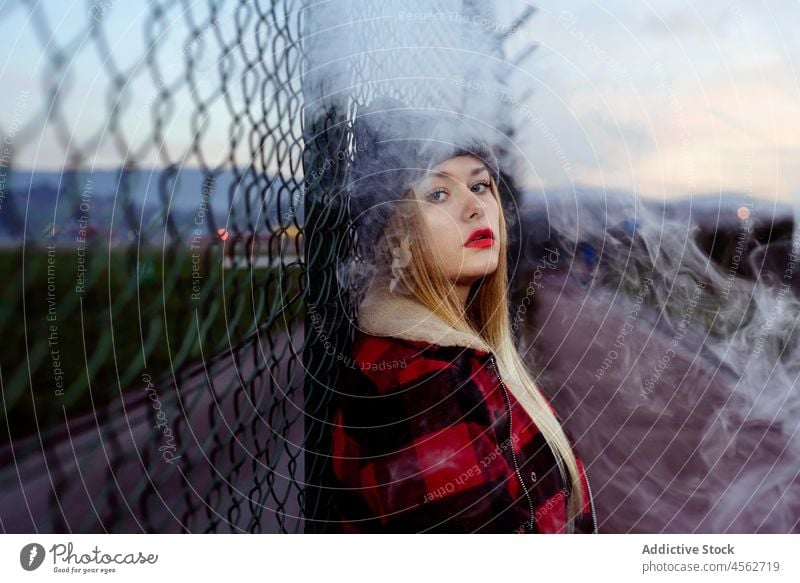 Young woman with wool hat leaning on the fence and smoking young portrait smoke looking at camera adult vaping alternative attractive beautiful blonde blow