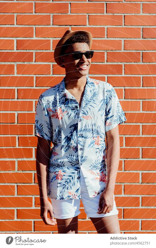Trendy young black man posing in the street ethnic teenager sunglasses african hat shirt floral american brick wall portrait standing smile look handsome