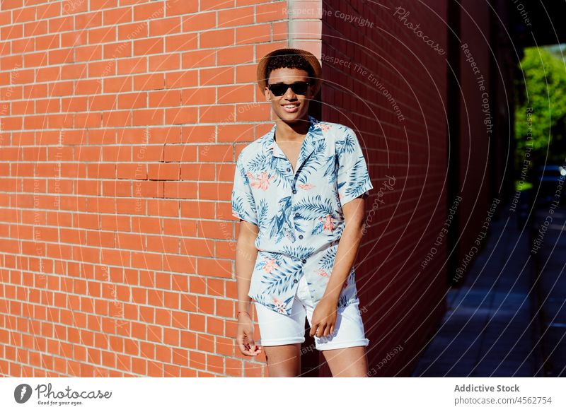 Trendy young black man posing in the street teenager sunglasses hat shirt floral brick wall portrait standing city urban look handsome attractive colorful