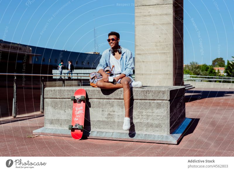 Young black man sitting on a bench using the phone male young mixed teenager skateboard school outdoors african urban street city backpack headphones scene