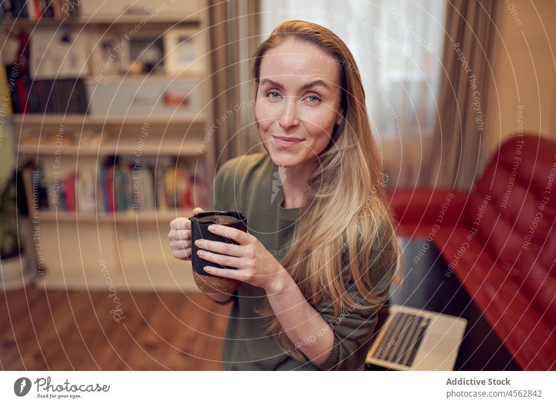 Cheerful woman with coffee in room hot drink living room domestic apartment couch laptop beverage home female sofa positive netbook lady light cup content