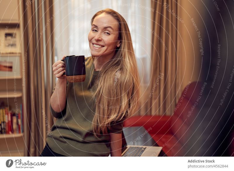 Smiling woman with coffee in room hot drink living room domestic apartment couch laptop beverage home female sofa positive netbook lady light cup content