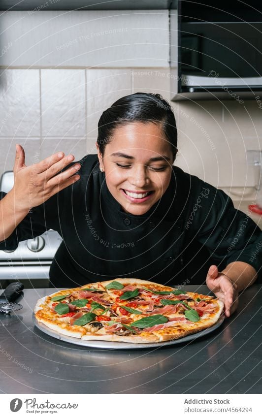 Smiling ethnic female chef in uniform enjoying pizza aroma woman meal cook smell sniff kitchen food tasty culinary dish restaurant work baked yummy appetizing