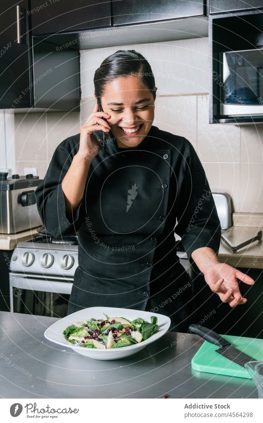 Smiling ethnic chef talking on smartphone with salad on table woman restaurant dish cook uniform culinary kitchen gadget job vegetable speak phone call meal