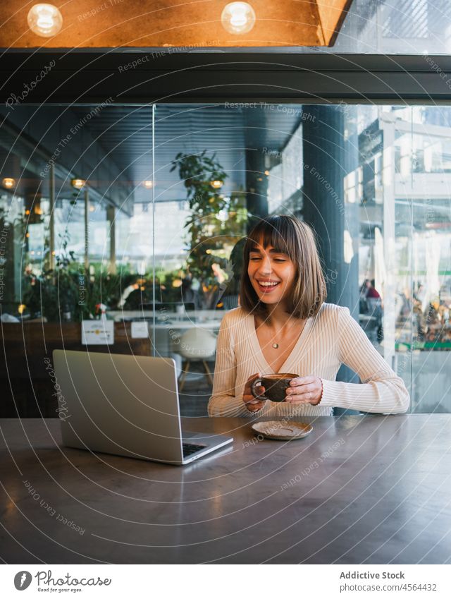 Cheerful woman with cup of coffee looking at laptop video call cafe using browsing device connection remote conversation freelance phone drink internet