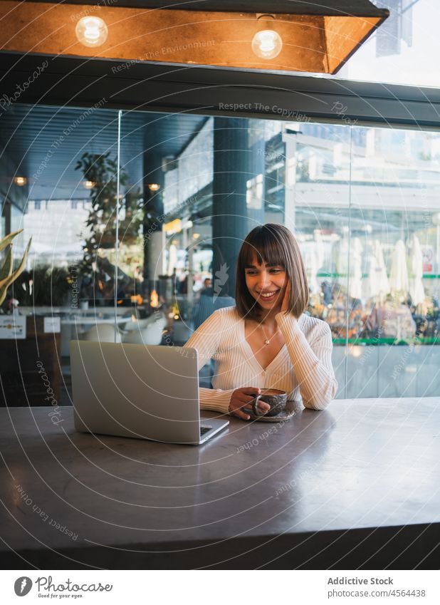 Cheerful woman with cup of coffee looking at laptop video call cafe using browsing device connection remote conversation freelance phone drink internet