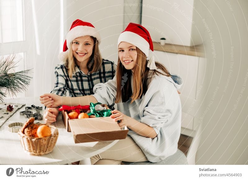 Two cute girls in Santa Claus hats are sitting at the kitchen table and packing sweet gifts to friends and relatives. A gift with your own hands, a sweet treat, festive preparation