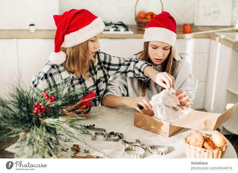 Two cute girls in Santa Claus hats are sitting at the kitchen table and packing sweet gifts to friends and relatives. A gift with your own hands, a sweet treat, festive preparation
