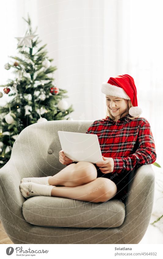 Happy teenage girl wearing santa claus hat using laptop in christmas room decorated with interiors chatting with friends and relatives new year hat santa