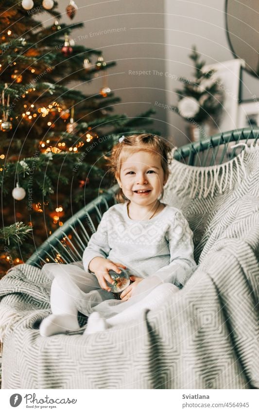 A little girl is sitting in an armchair and smiling against the background of a Christmas tree on New Year's Eve. Christmas mood, expectation of magic eve