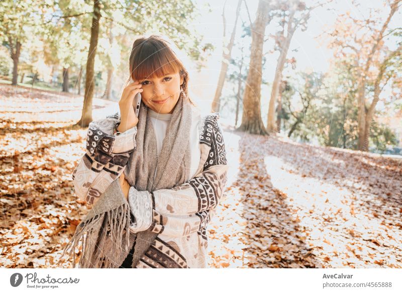 Happy girl using smart phone during Autumn in a park taking selfie, woman talking on mobile in fall. Video call outdoors, receiving good news. person female