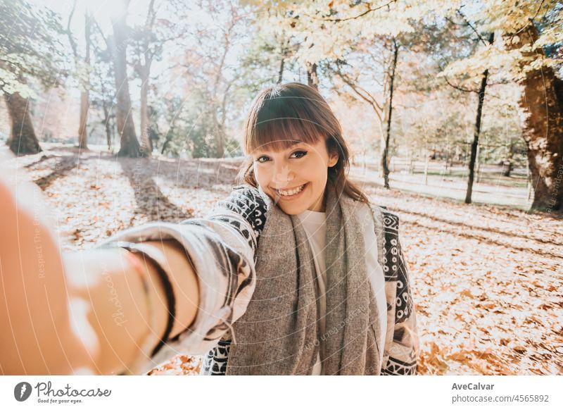Happy girl using smart phone during Autumn in a park taking selfie, woman talking on mobile in fall. Video call outdoors, receiving good news. person female