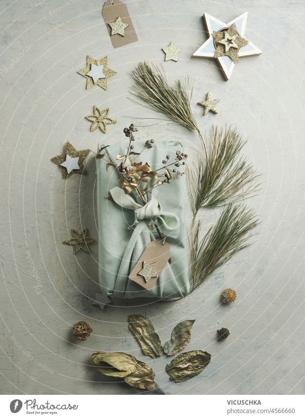 Eco-friendly Christmas gift,  wrapped in fabric with golden decoration stars, branches on pale grey table. Sustainable zero waste gift packaging. Top view.