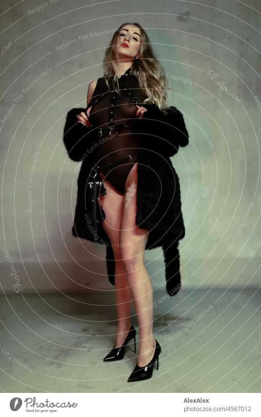 Portrait of a young blonde dominatrix in high heels in front of a concrete wall. She wears black underwear and and a black short coat Woman Young woman pretty
