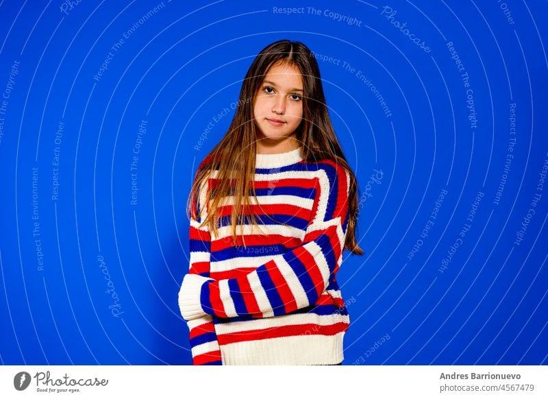 Adorable eleven years old girl wearing a red and blue striped sweater on blue studio background, pretty brown-haired fringe hairstyle european appearance child pose indoor smiling look at camera, generation Z concept