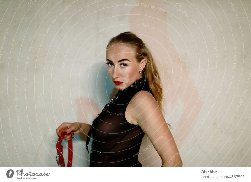 Portrait of a young blonde dominatrix in front of a concrete wall. She wears black underwear and holds a red leather collar promptly in her hand and looks very serious into the camera