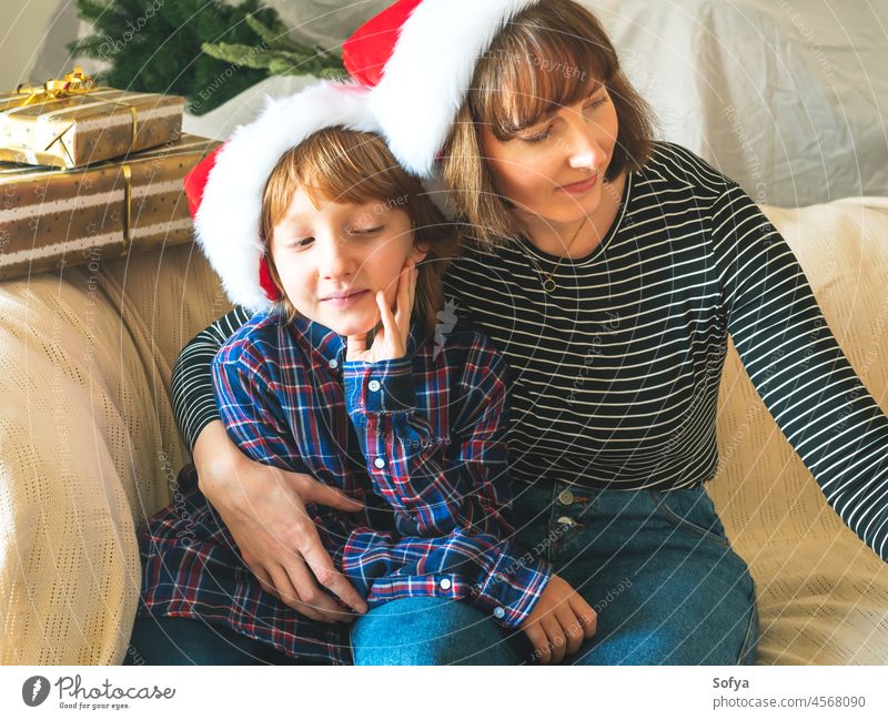 Mother and son in santa claus caps on couch, christmas mother lifestyle kid child family little december casual new year together hugging presents tree morning