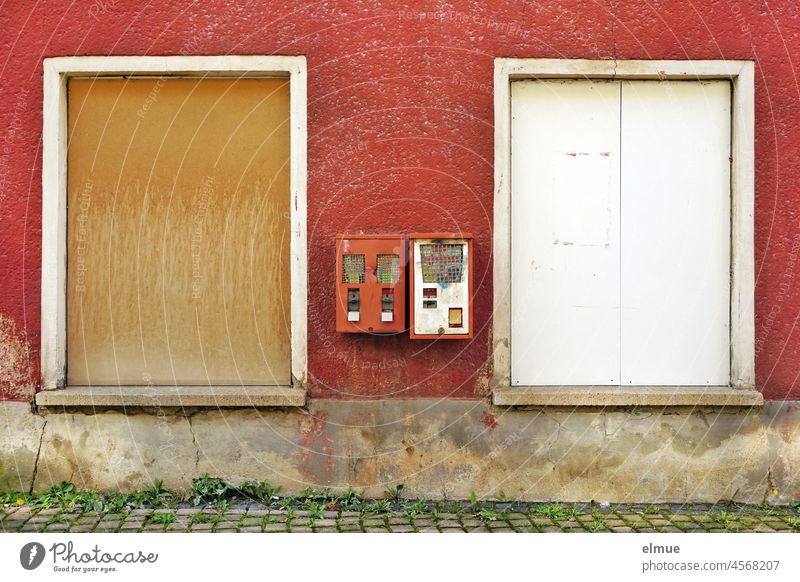 Two old, defective chewing gum vending machines / vending machines on a red plastered house wall between two windows boarded up with wood Gumball machine