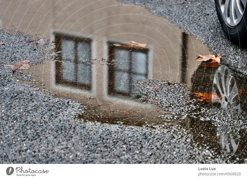 Parts of a building and the wheel of a parked car are reflected in a puddle at the side of the road. Street Asphalt Wet Puddle reflection