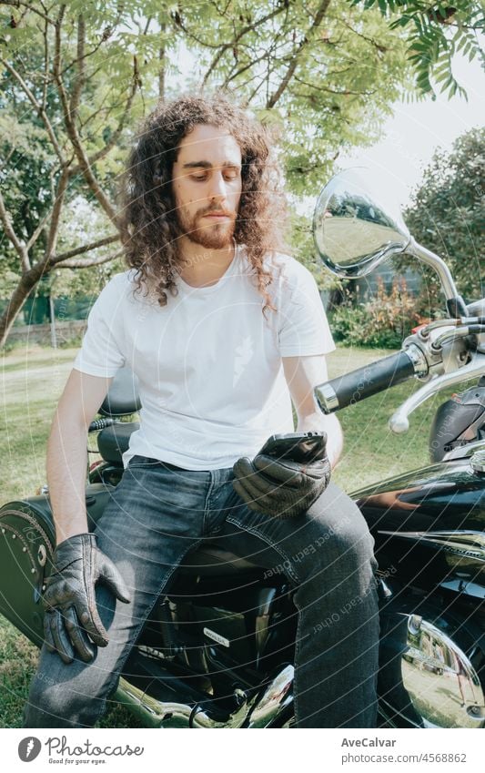 Young long hair motorbike guy checking his phone while sitting on his old school motorbike during a break from the road route. Liberty life, young man heavy metal, white tshirt and gloves.