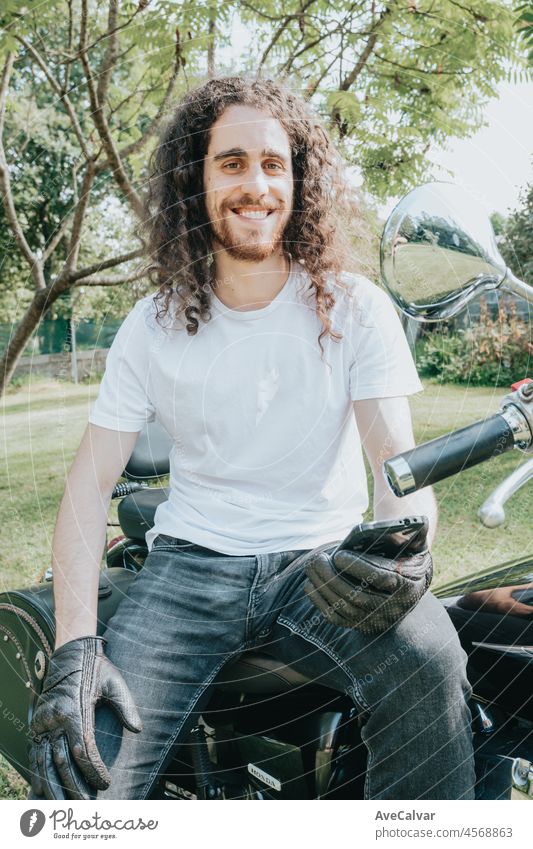 Young long hair motorbike guy smiling to camera while checking his phone while sitting on his old school motorbike during a break from the road route. Liberty life, young man heavy metal, white tshirt