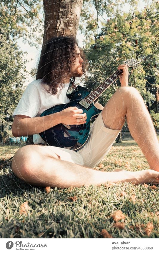 Young long hair hippie man smiling playing guitar in park while resting against a tree. Hobby art activity for young adults. Authentic manhood moment. person