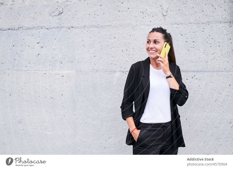 Smiling lady speaking on smartphone against wall woman cheerful smile cellphone talk phone call conversation using street female gadget formal hand in pocket