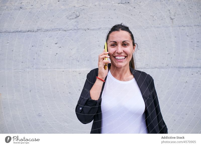 Smiling lady speaking on smartphone against wall woman cheerful smile cellphone talk phone call conversation using street female gadget formal worker lifestyle