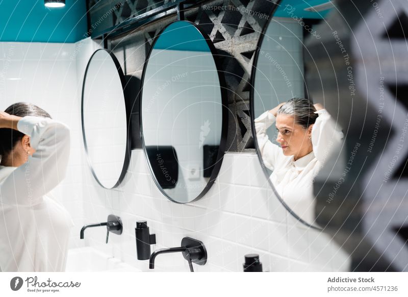 Woman fixing hair near mirror in restroom woman hairdo ponytail reflection style modern hairstyle sink female clean outfit worker elegant care tap contemporary