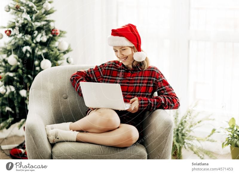 Happy teenage girl wearing santa claus hat using laptop in christmas room decorated with interiors chatting with friends and relatives new year hat santa