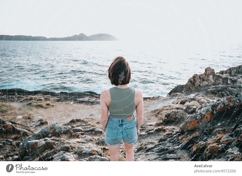 A young woman walks on the path of rocks on the shore and looks at the evening sea.Young hipster girl doing some hiking on the shore, traveling and low budget travel concepts, copy space